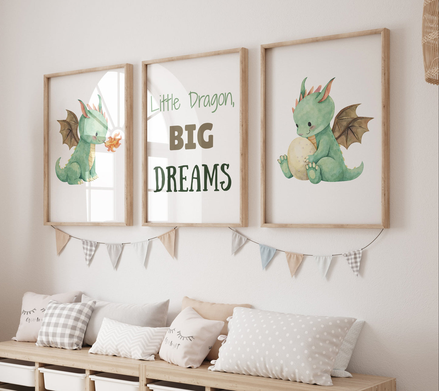 A styled children's room corner featuring three framed wall art prints with dragon illustrations and the text 'Little Dragon, BIG DREAMS' above a light wood storage bench with pillows. A tall woven giraffe toy stands on the left.