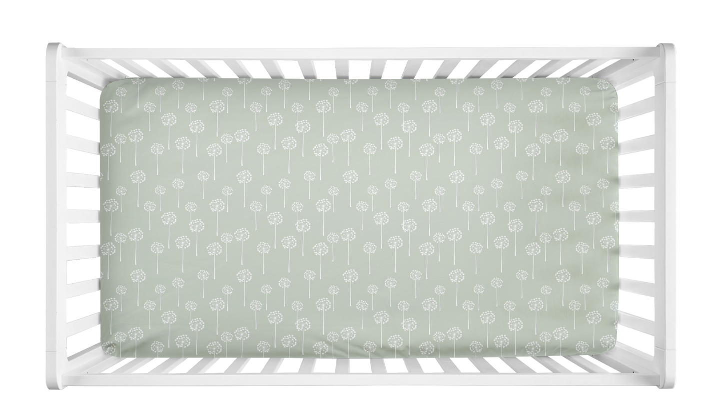 Crib sheet with sage green background and white dandelion print, styled in a white crib for a botanical or minimalist baby girl nursery