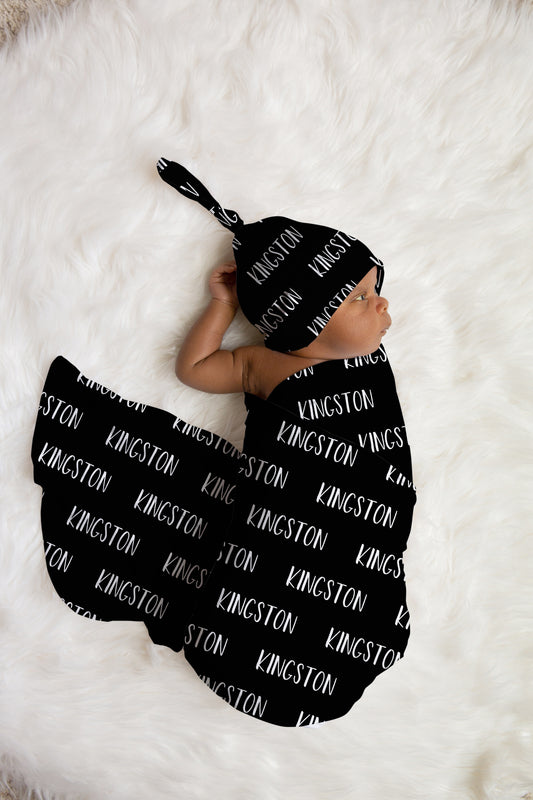 Newborn baby wrapped in personalized black swaddle and matching name hat